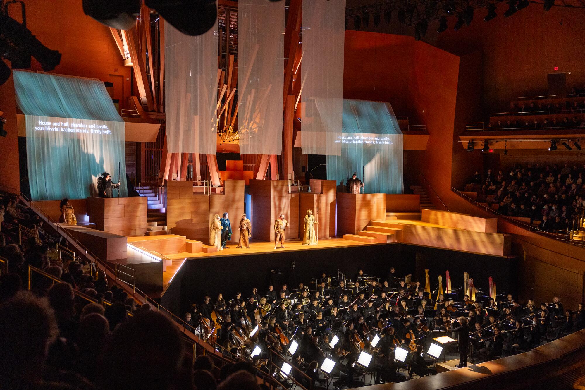 Singers stand on a golden lighted stage with screens behind them and a full orchestra below.