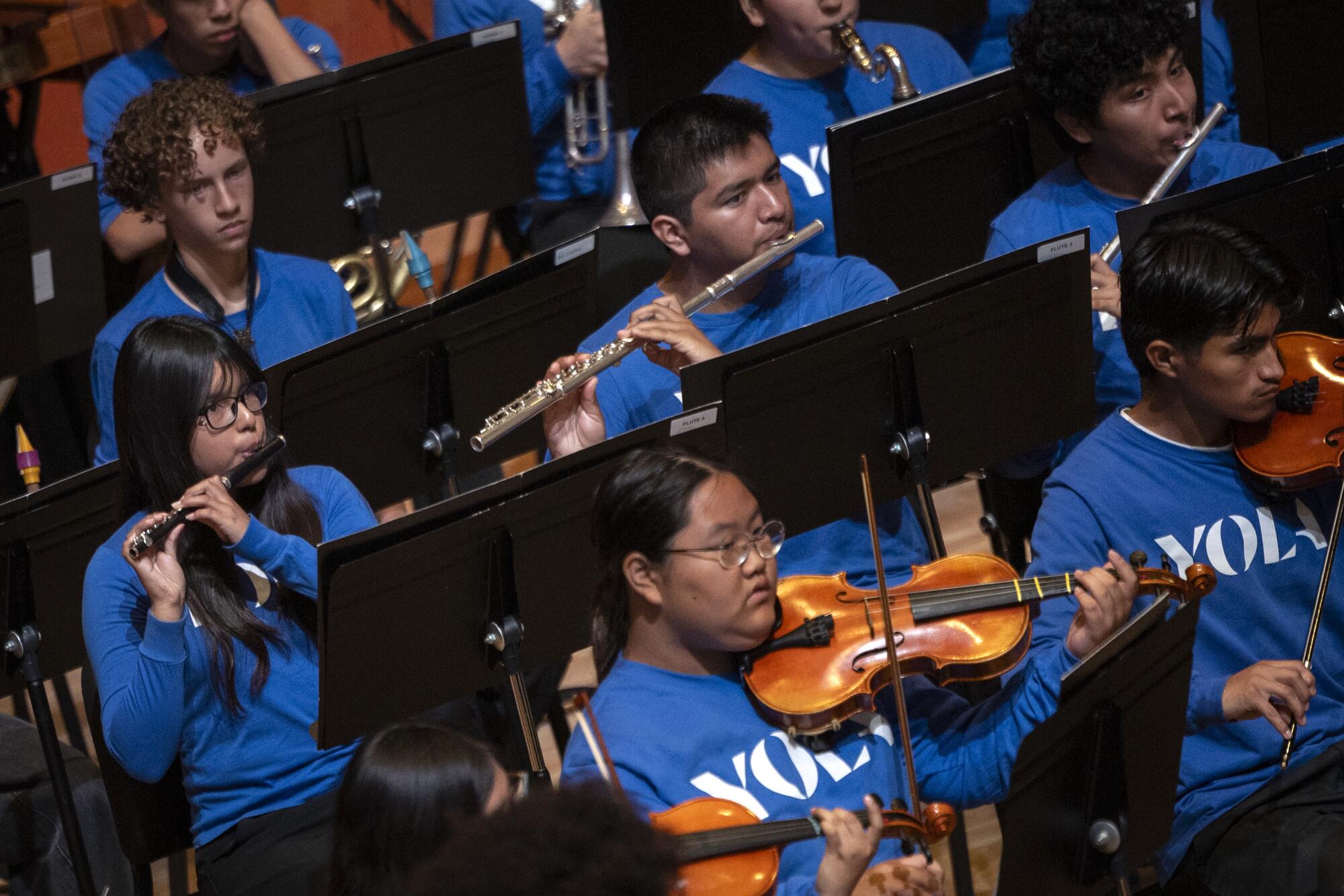 Children, wearing blue YOLA shirts, play instruments in an orchestra.