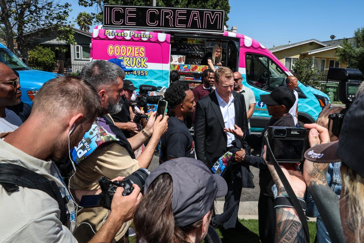 A crowd uses cellphones and cameras to record a man in a dark suit walking in front of a red, white and blue food truck