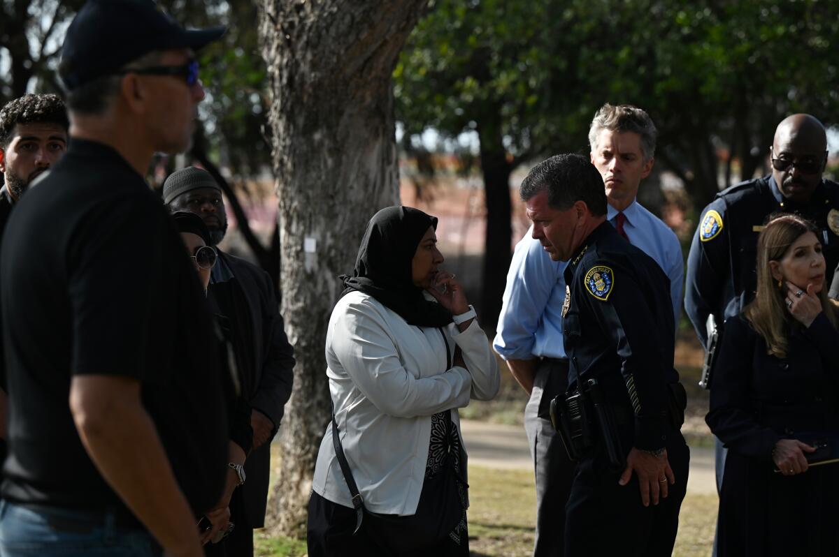 A man in dark police uniform, right, speaks to a woman wearing a black headscarf, with other people standing nearby