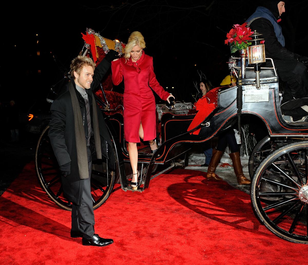 A man in a dark suit standing near a blond woman in a red dress stepping off a carriage on a red carpet 