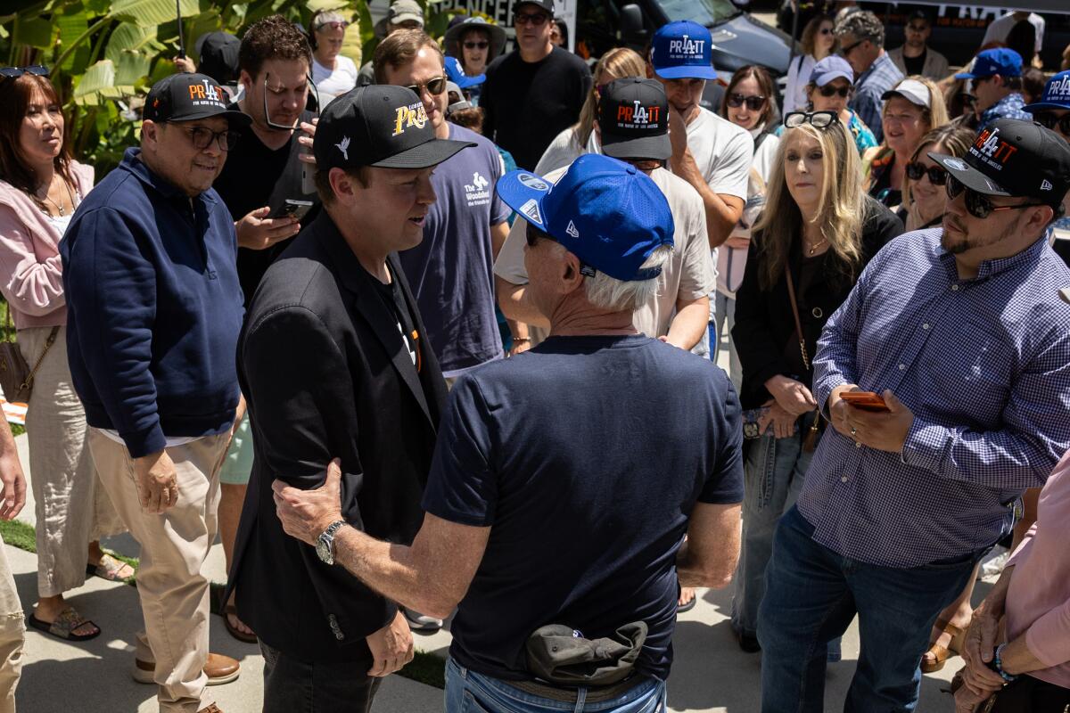 A man in a blue ball cap holds the arm of another man, in a dark suit and dark ball cap, in a crowd