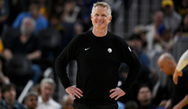 A man wearing a black Golden State Warriors sweatshirt stands with hands on hips, smiling slightly, with a blurred crowd behind him.