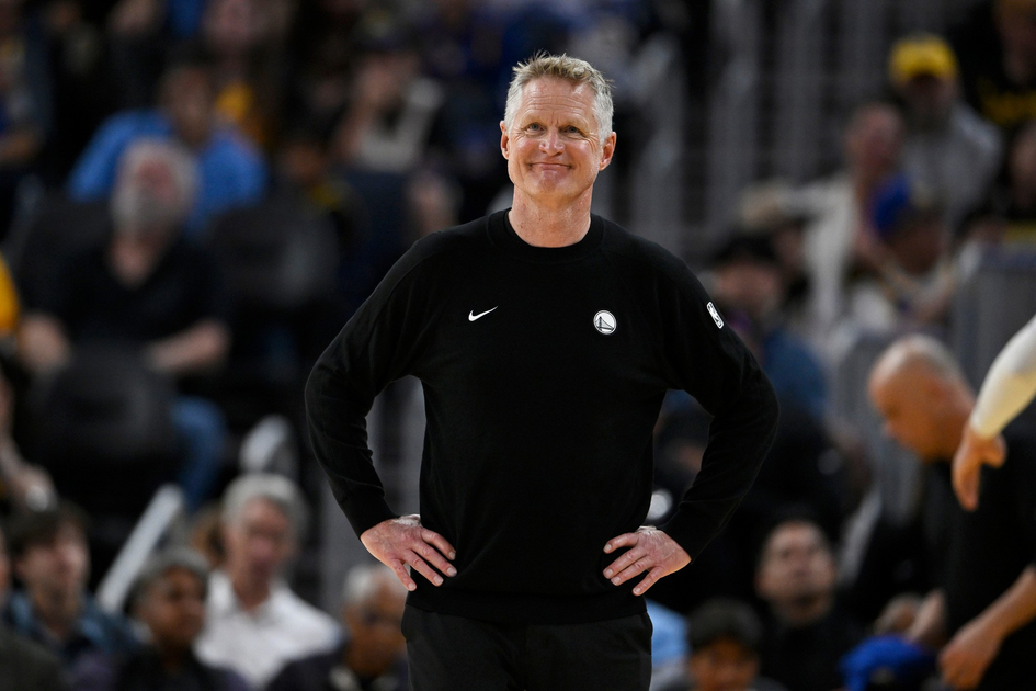 A man wearing a black Golden State Warriors sweatshirt stands with hands on hips, smiling slightly, with a blurred crowd behind him.