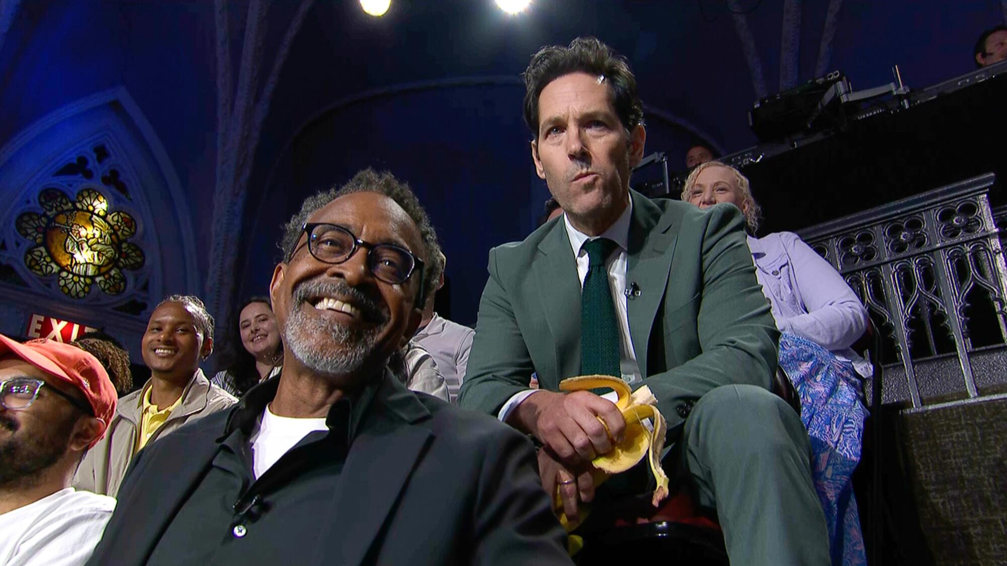 Two men seated in the audience with other guests on risers.