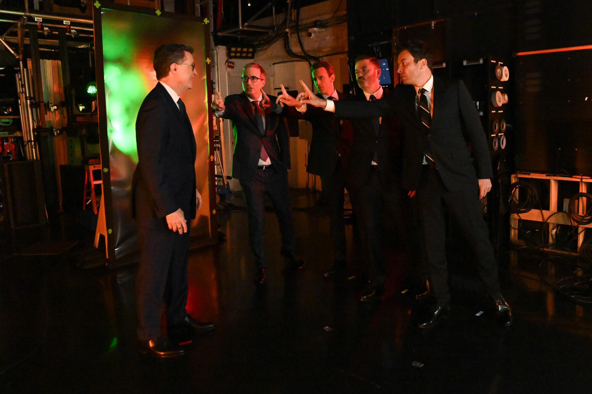 A man stands facing four men who are holding out their arms with fingers pointed at his face.