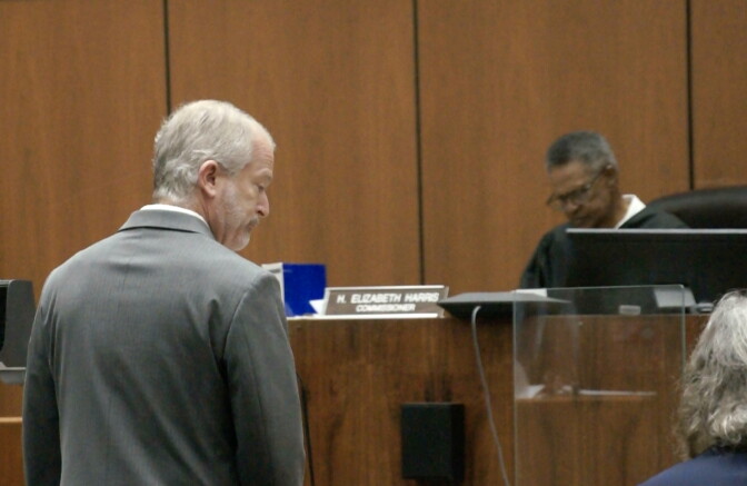 A man in a grey suit jacket stands looking down in a courtroom, as a judge in the background looks down as well.
