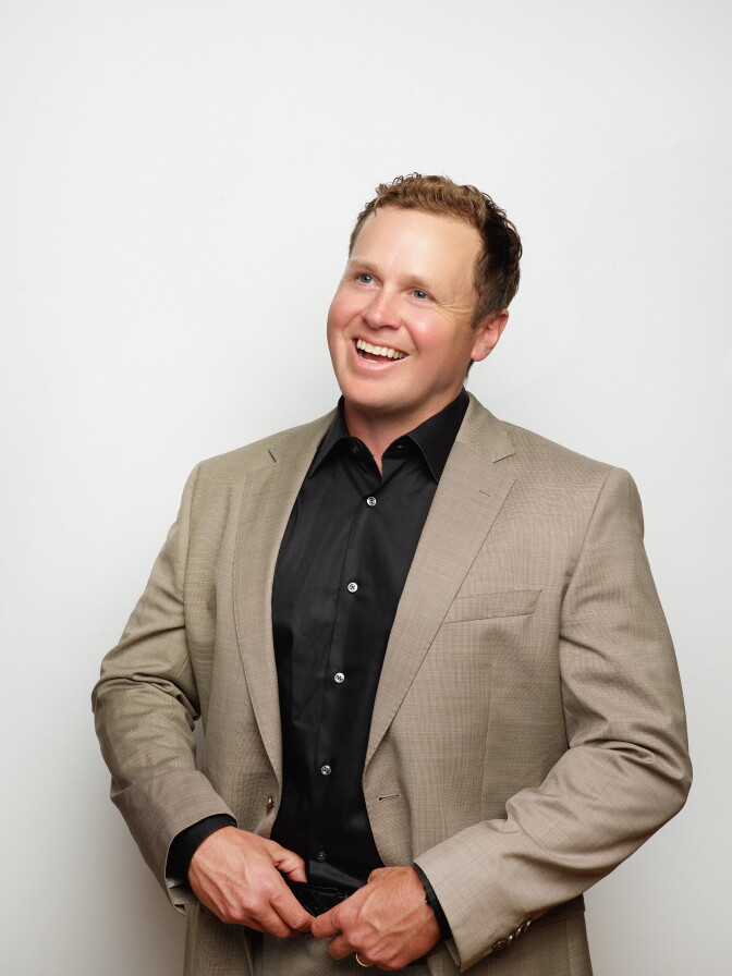 A white man with reddish-brown hair wears a tan suit with a black shirt and smiles.