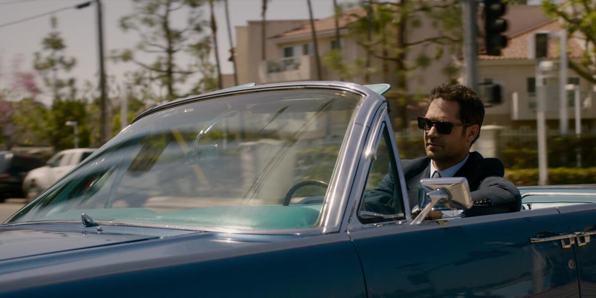A man in a suit and sunglasses driving a blue convertible in Los Angeles.