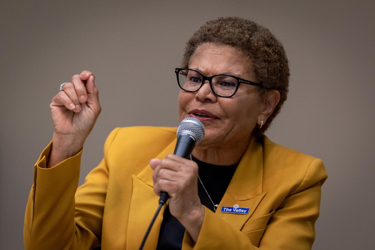 A woman with curly, short brown hair, wearing glasses and a mustard yellow jacket, speaks while holding a microphone