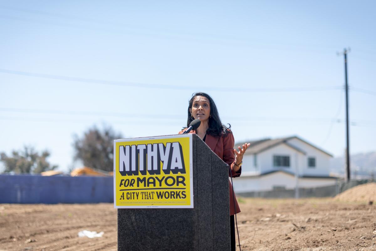 A woman speaks at a lectern with a sign that says 'Nithya for Mayor' in a dirt lot