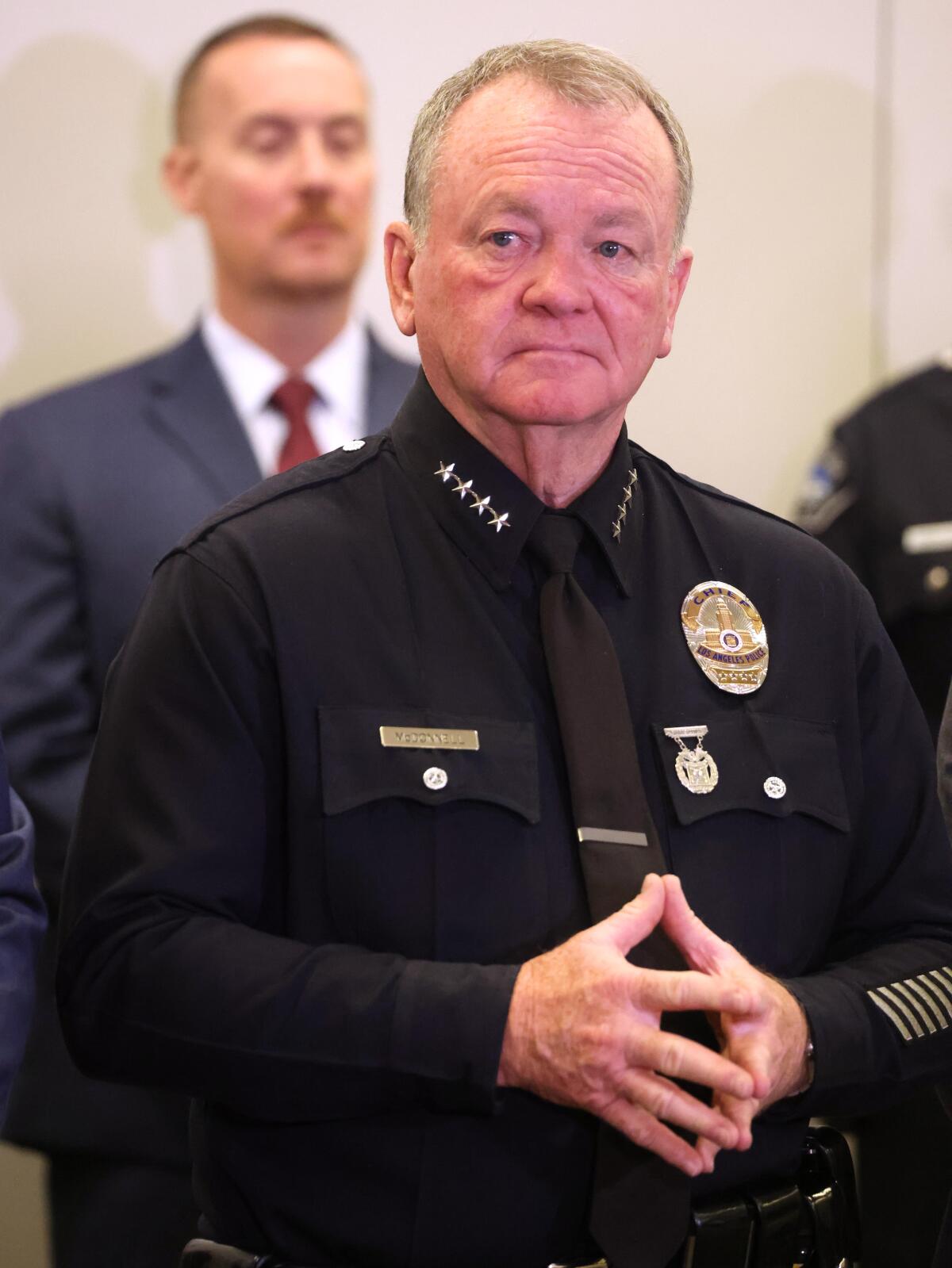 A man in dark police uniform holds his fingers together as he stands in front of another man