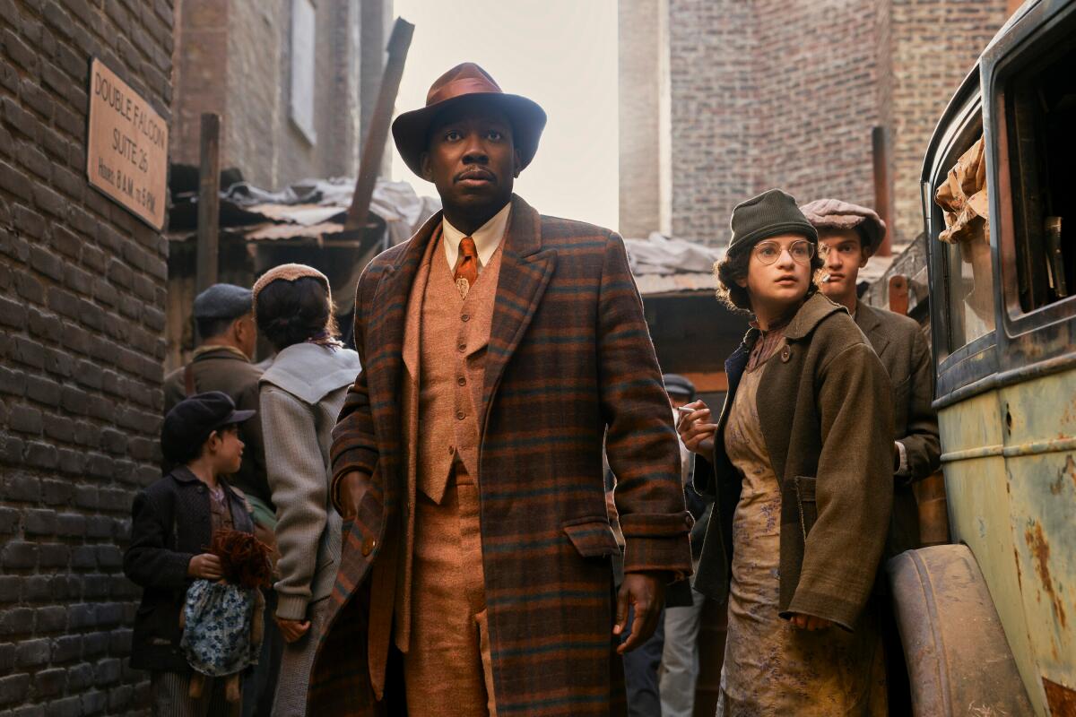 A man in a plaid coat, orange-hued suit and brown hat stands in an alleyway crowded with people.