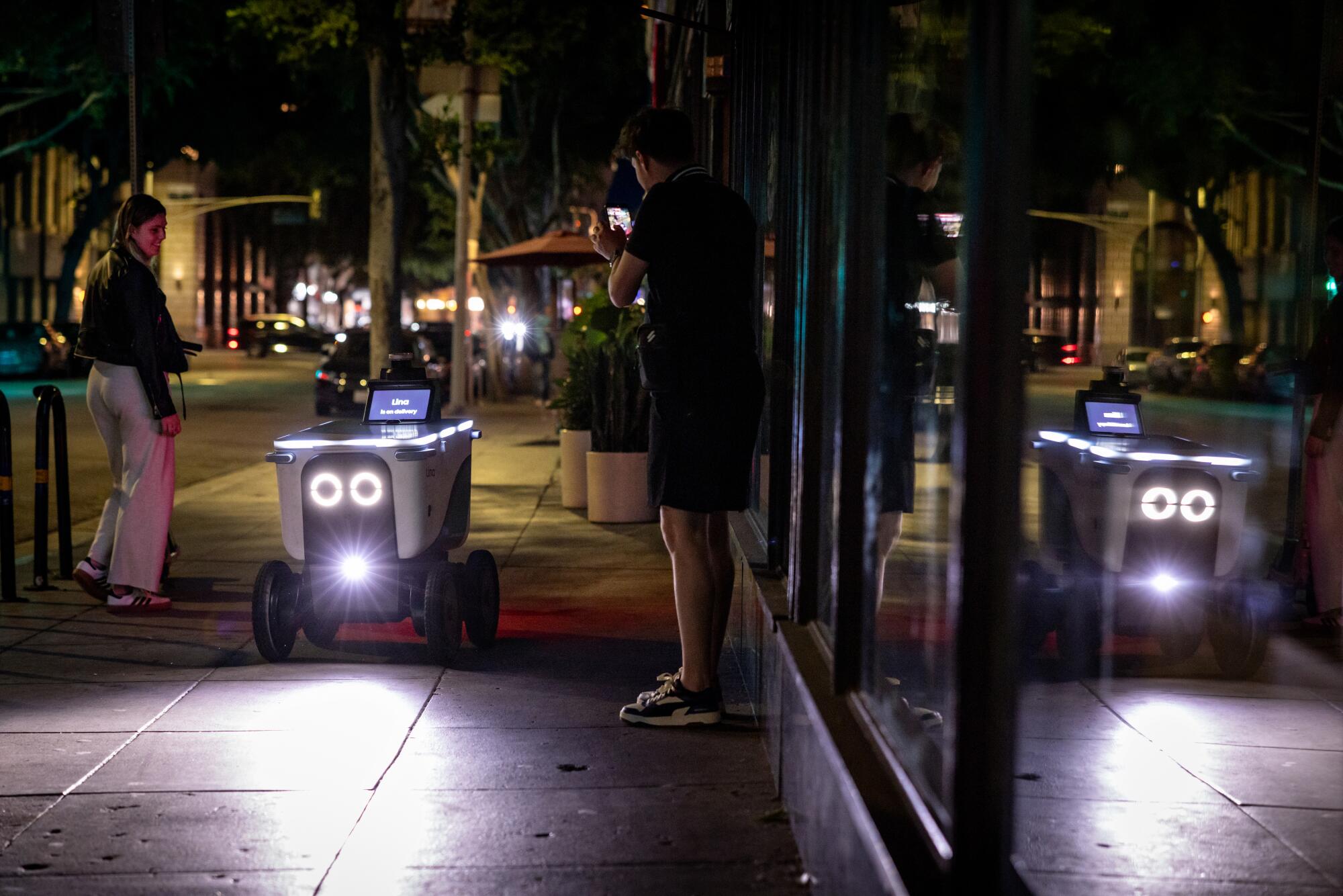 A food delivery robot with built-in headlights navigates near 8th and Olive streets in downtown Los Angeles.