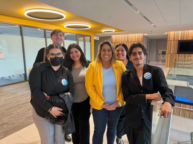 A group of five young people standing around a woman with a bright yellow blazer in an office building.