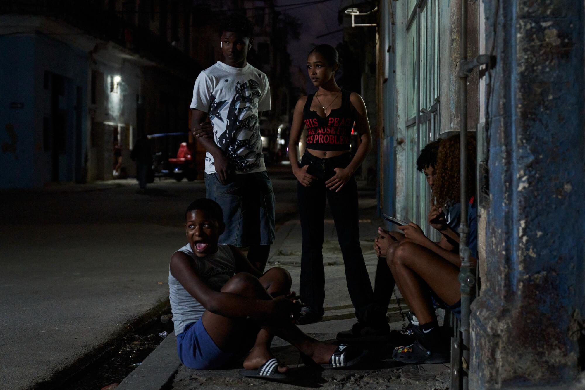 Two young people standing and two others sitting on the ground and on the steps of a building next to a darkened road