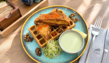 Two pieces of breaded fried chicken on top of a waffle, served with a creamy dipping sauce, sprouts, lemon wedge, and candied walnuts on a blue plate.