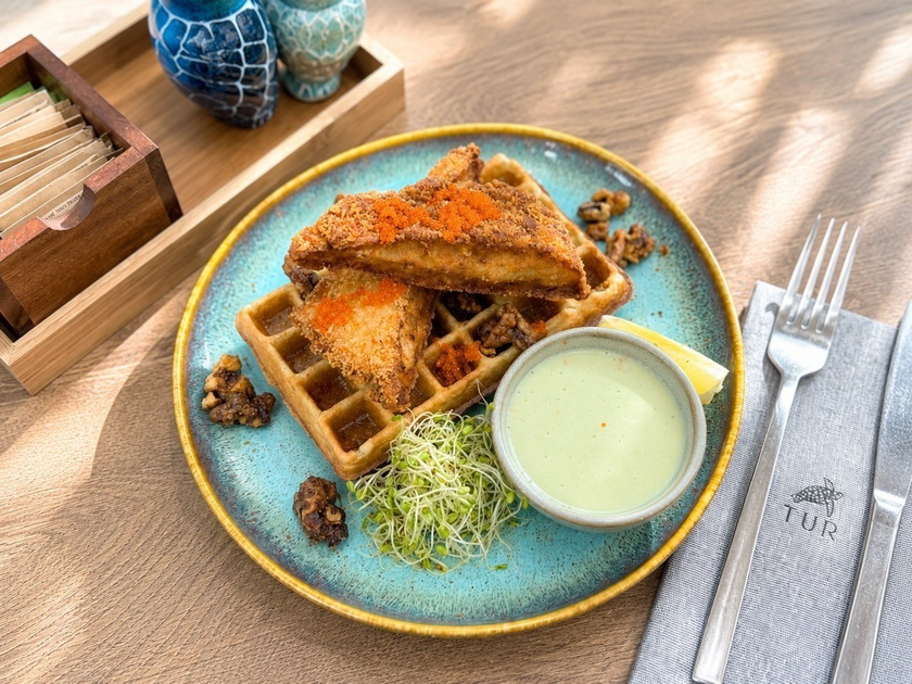 Two pieces of breaded fried chicken on top of a waffle, served with a creamy dipping sauce, sprouts, lemon wedge, and candied walnuts on a blue plate.