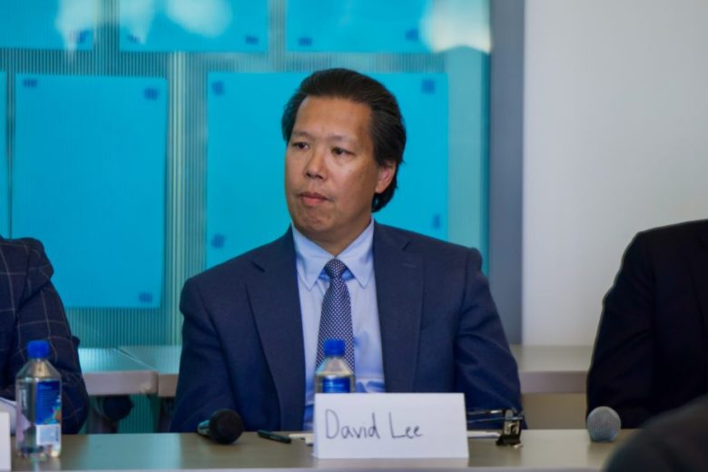 A man in a blue suit sits at a table with a name card reading "David Lee," a microphone, and water bottles in front of him.