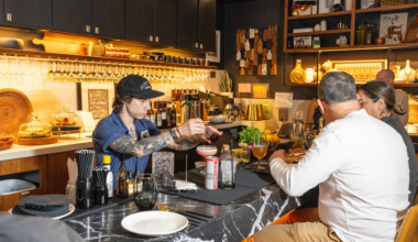 A tattooed bartender in a blue shirt and black cap prepares drinks behind a marble bar counter while two seated customers eat and chat.