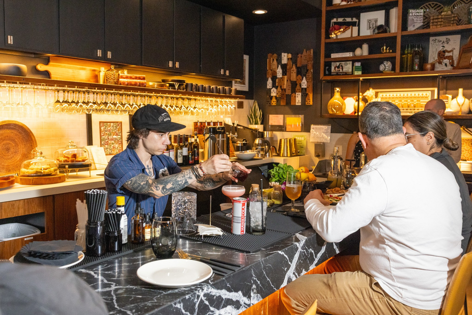 A tattooed bartender in a blue shirt and black cap prepares drinks behind a marble bar counter while two seated customers eat and chat.