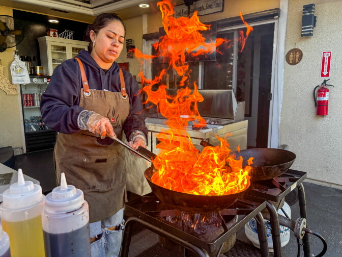 A woman in a brown Lomo Fuego apron stirs a wok over a powerful outdoor burner, producing dramatic flames that leap several feet into the air in a backyard restaurant's patio area.