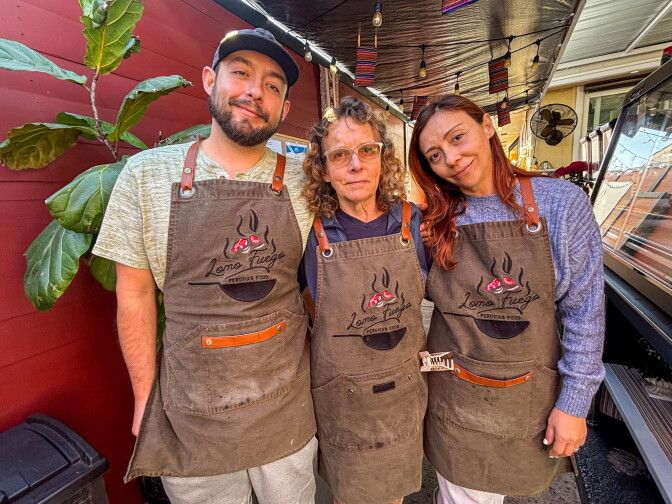 Three people with medium-light skin tones wearing matching brown Lomo Fuego aprons stand together, smiling, in the restaurant's covered backyard dining area. String lights and colorful Peruvian textiles hang overhead.