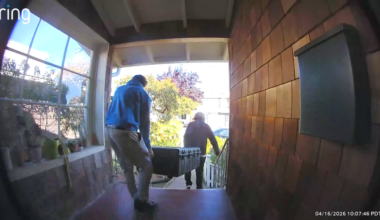 Two people carry a large black case up the porch steps of a house, with plants on the left window sill and a mailbox on the right wall.