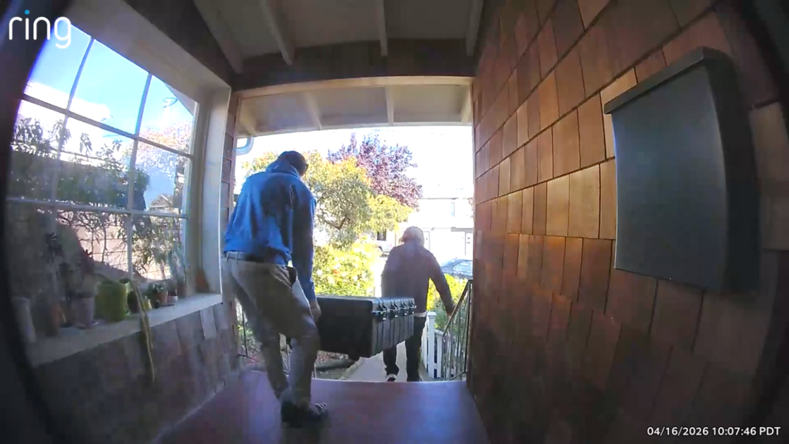 Two people carry a large black case up the porch steps of a house, with plants on the left window sill and a mailbox on the right wall.