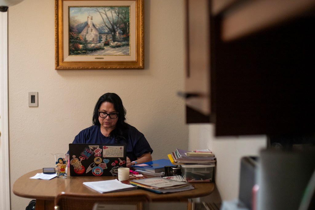 A person wearing a blue shirt and glasses is working on a laptop at a kitchen table, with books and school materials scattered around, in the corner of a kitchen in a home. In the background, a framed painting hangs directly above the person as they work, while in the foreground is a view of a kitchen cabinet.
