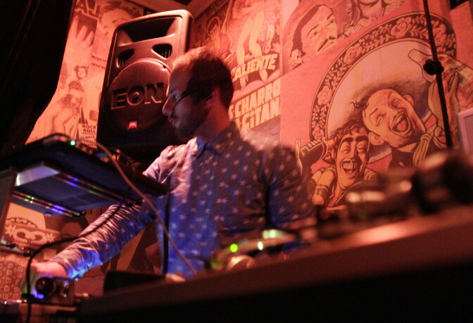 A light-skinned man wearing a blue and white polka dot button up stands behind a DJ set. A large speaker is set up behind him.