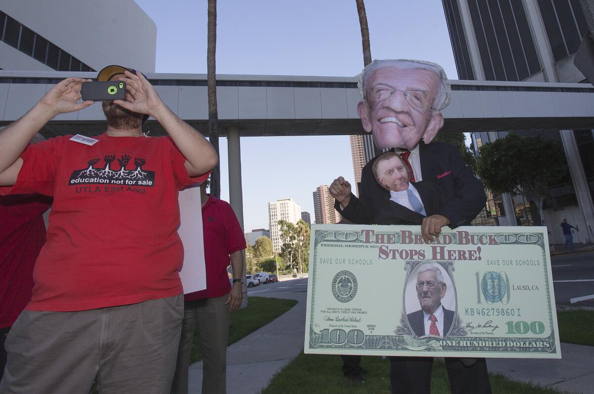 A man holds a camera in front of his face next to a person in a giant puppet head and holding a big fake $100 bill.