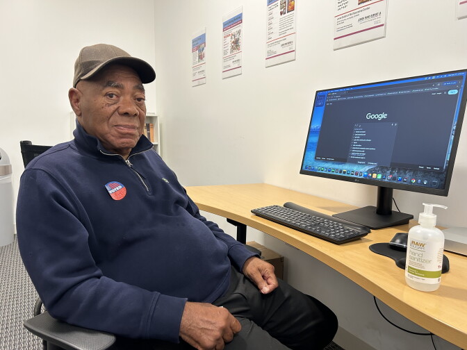 A Black man wearing a tan baseball cap, blue quarter zip sweater and black pants is sitting in front of a computer with a webpage open to Google. He has an "I voted" blue and red sticker affixed to his chest.