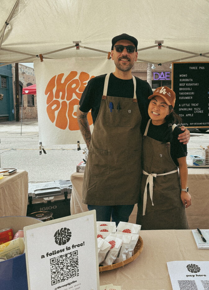 A light skinned man wearing a baseball hat, dark glasses and a chef's apron, has his arm around a smiling Asian woman also wearing a baseball cap and a chefs apron. They are standing inside a pop up tent surrounded by kitchen equipment.
