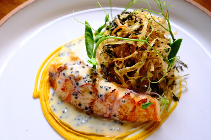 A white plate holds a beautifully laid out dish, with crispy seaweed and lobster tail on a cream colored sauce, surrounded by a yellow swirl.