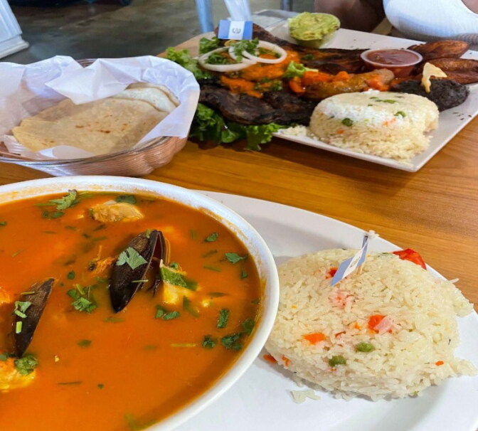 A borth with seafood in it. A side of rice and tortiallas next to another plate of grilled meats and rice.