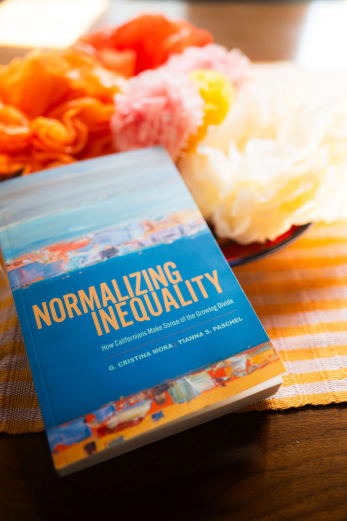 a book with a blue cover placed on a table with paper flowers. The title is Normalizing Inequality: How Californians Make Sense of the Growing Divide