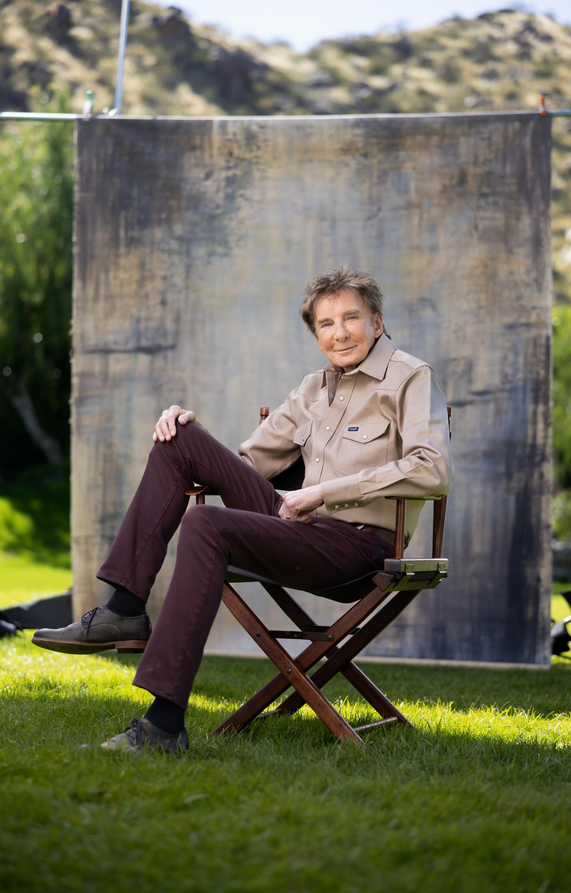 Barry Manilow, wearing a khaki shirt and brown pants, sits on a chair on his lawn.