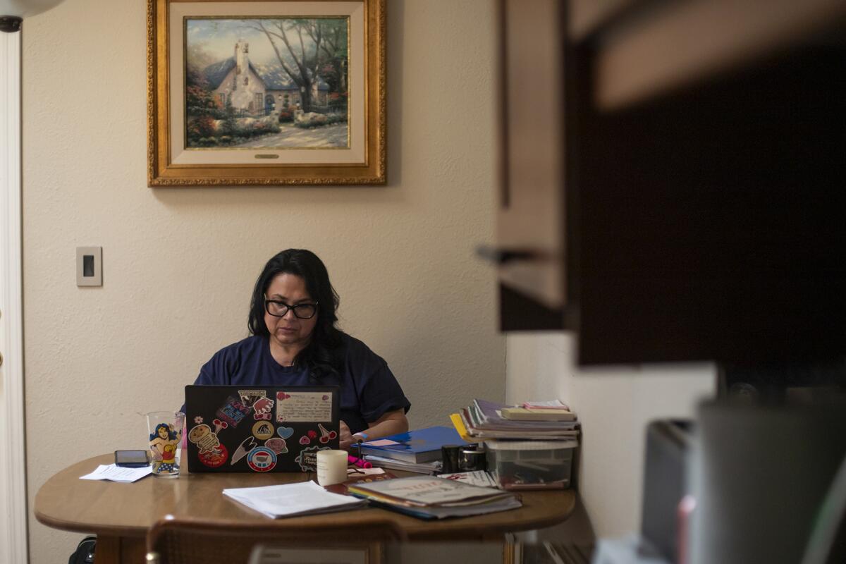 A person wearing a blue shirt and glasses works on a laptop at a kitchen table, with books and school materials scattered.
