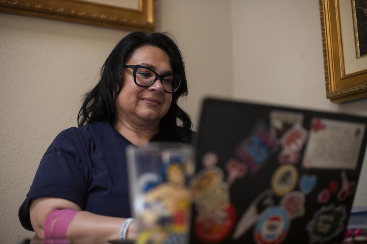A close-up shot of a person wearing a blue shirt and glasses working on a laptop on a kitchen table inside a house. In the foreground, out of focus, is the back of the laptop with decorative stickers and a colorful glass of water as the person types on the keyboard.
