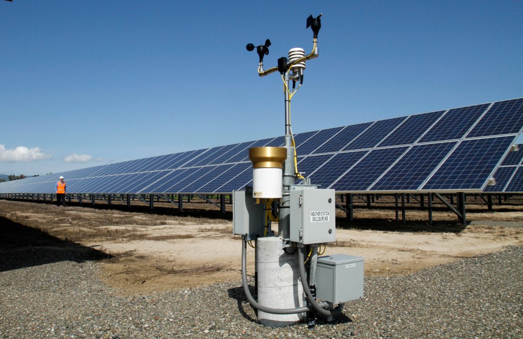 A weather station stands in front of solar panels at a solar energy site.