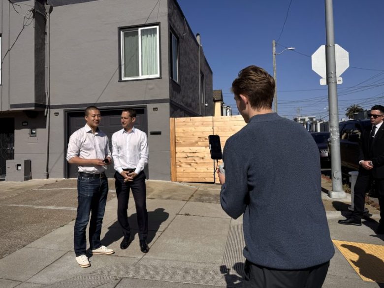 Three men stand on a residential street; two pose in front of a house while the third records them with a smartphone on a stabilizer. A fourth man stands nearby wearing sunglasses.