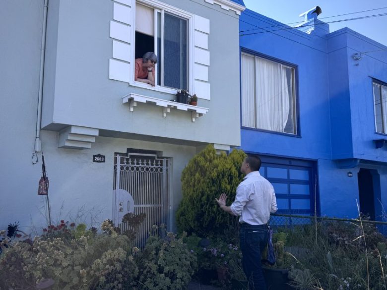 A man stands outside a light blue house speaking to an older woman who is leaning out of an upstairs window.
