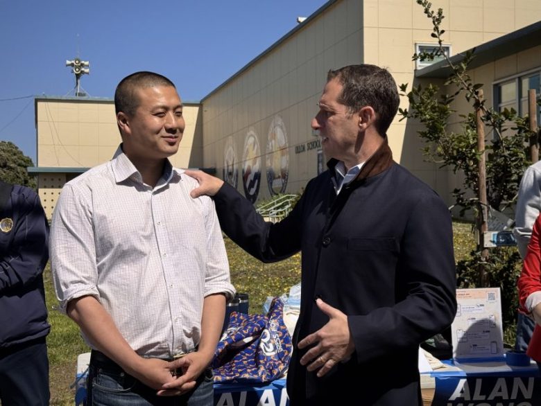 Two men stand outside near a table with items; one man rests his hand on the other's shoulder as they talk in front of a beige building with murals.
