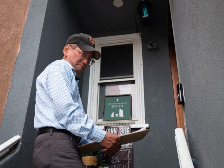 An older man wearing a cap and glasses holds a clipboard while standing in front of a gray building's entrance, looking back over his shoulder.
