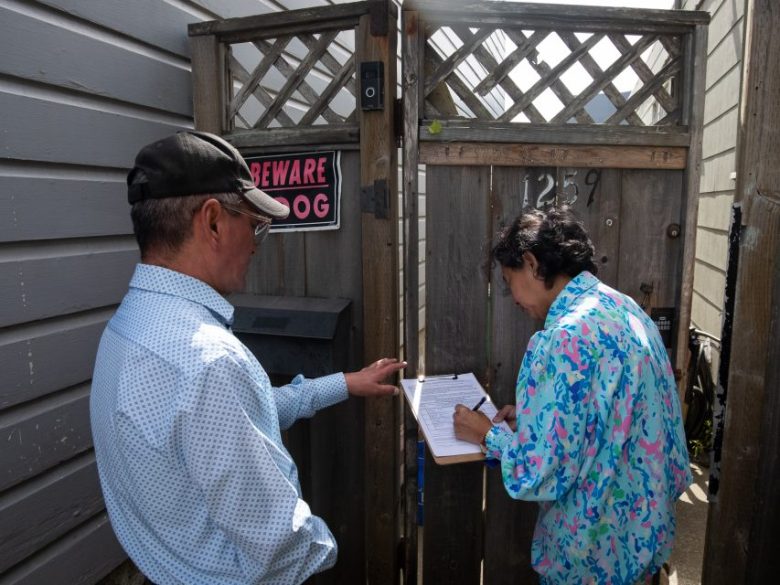 A man stands near a gate with a "Beware of Dog" sign as a woman writes on a clipboard outside a house numbered 1259.