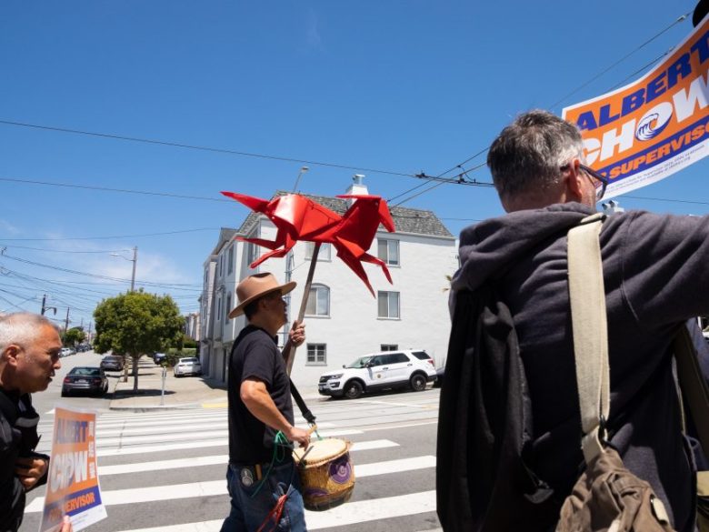 A man holding a red origami dragon and playing a drum walks across a street during a campaign event, with others holding political signs nearby.