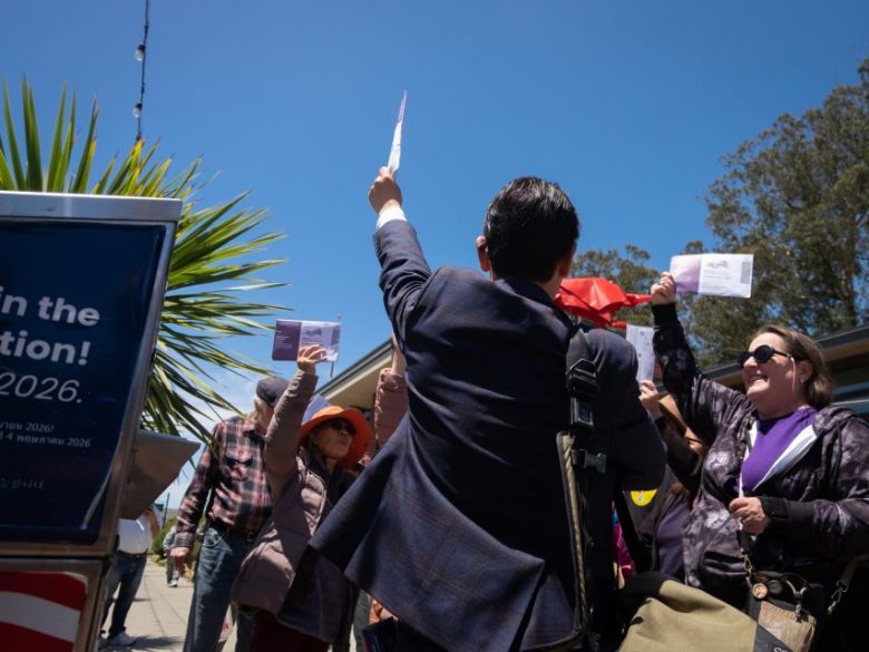 A group of people outdoors hold up papers while a man in a suit faces them and raises a document. The sky is clear and sunny.