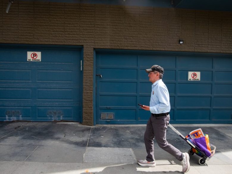 A man walks on a sidewalk pulling a small cart with groceries past blue garage doors marked with "No Parking" signs.