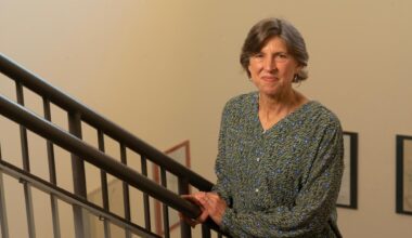 Woman in green patterned blouse on indoor staircase, holding railing and smiling towards the camera.